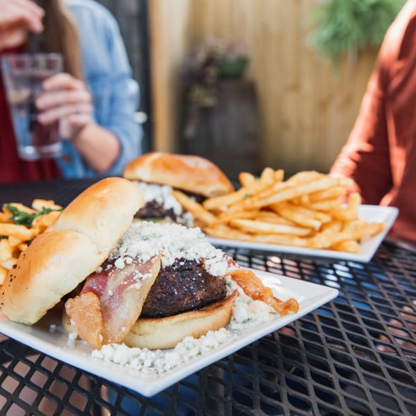 Two gourmet burgers with bacon, cheese, and fries on plates at an outdoor restaurant table.