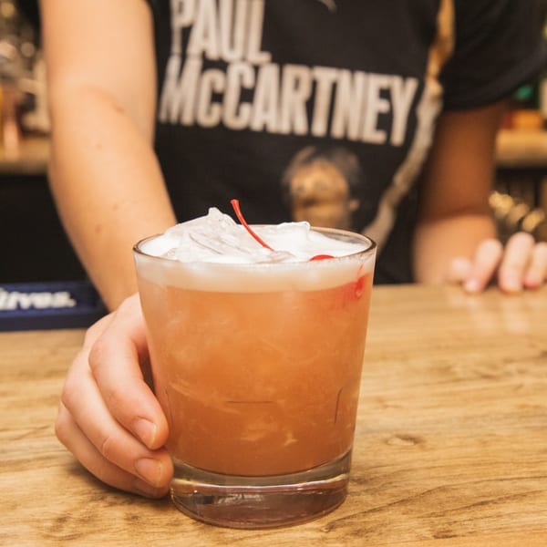 Bartender's hand presenting a cocktail with ice and a cherry on a wooden bar.