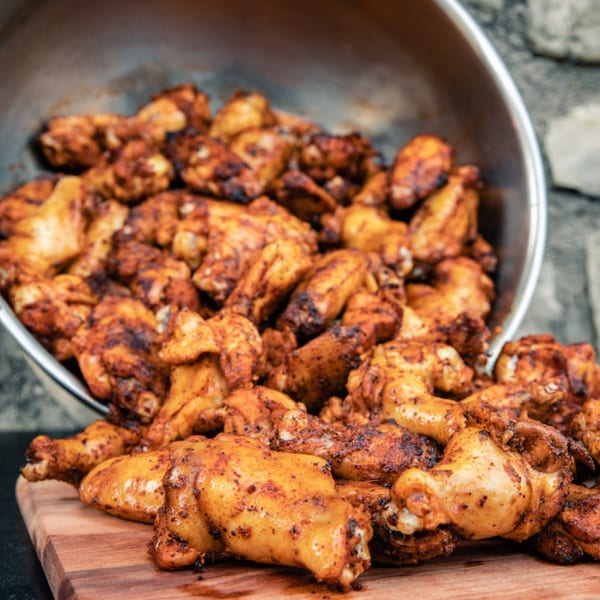 A large metal bowl overflowing with seasoned, cooked chicken wings onto a wooden cutting board.