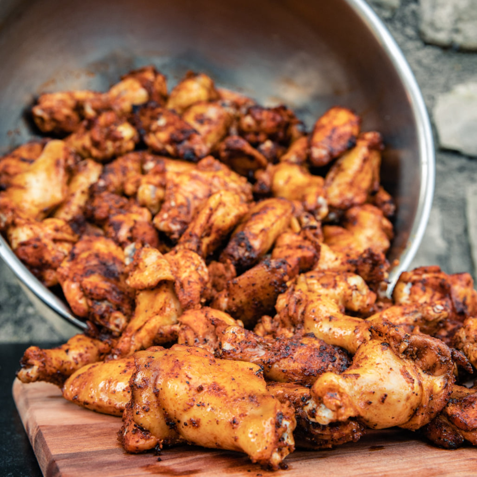 A large metal bowl overflowing with seasoned, cooked chicken wings onto a wooden cutting board.