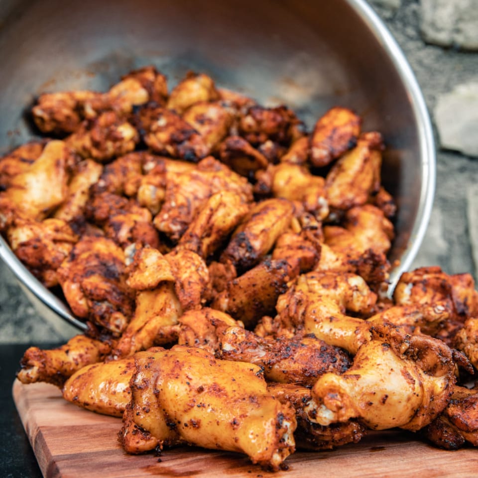 A large metal bowl overflowing with seasoned, cooked chicken wings onto a wooden cutting board.