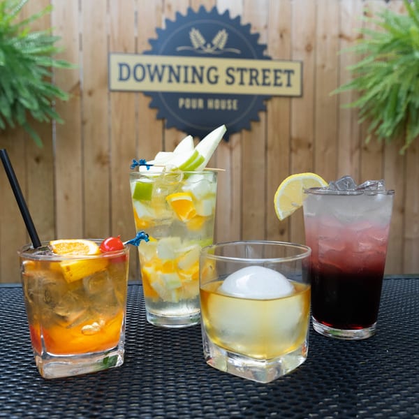 Four colorful cocktails on a black mesh table with a wooden fence and "Downing Street Pour House" sign in the background.