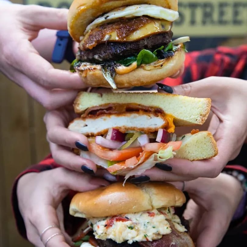 Three hands holding a towering stack of three different gourmet burgers with various toppings.
