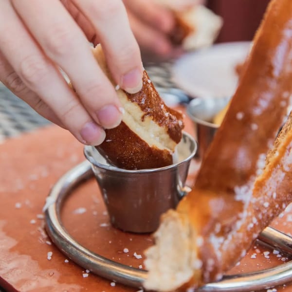 A hand dips a piece of a large, salted pretzel into a small metal cup of dipping sauce.
