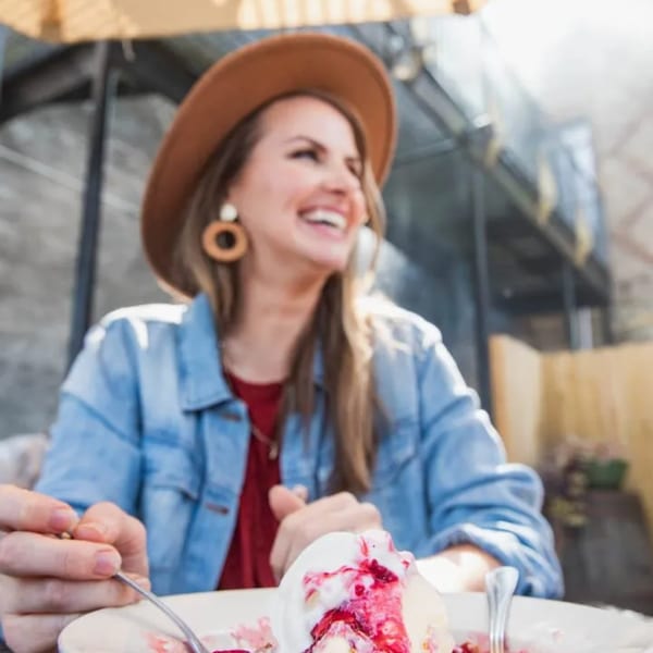 A smiling woman in a hat and denim jacket enjoys a dessert with berries and cream.