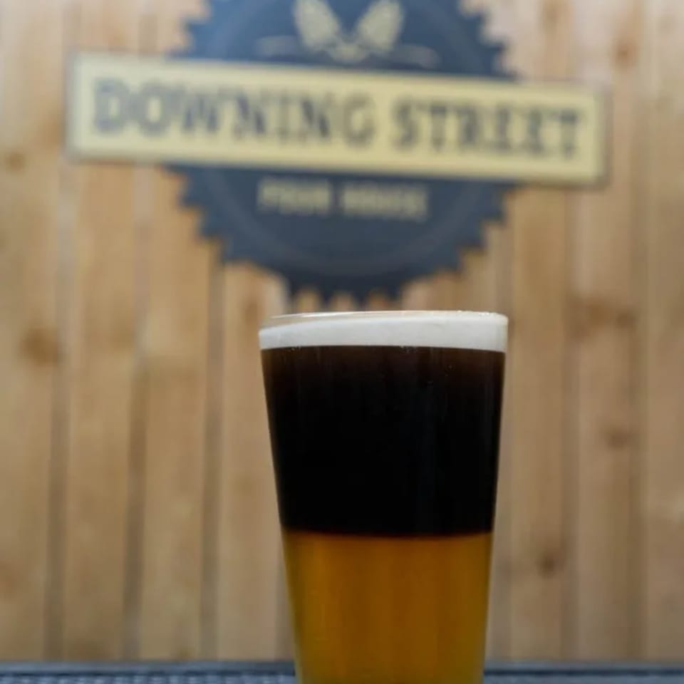 A glass of dark beer with a golden bottom layer, topped with white foam, in front of a wooden wall.