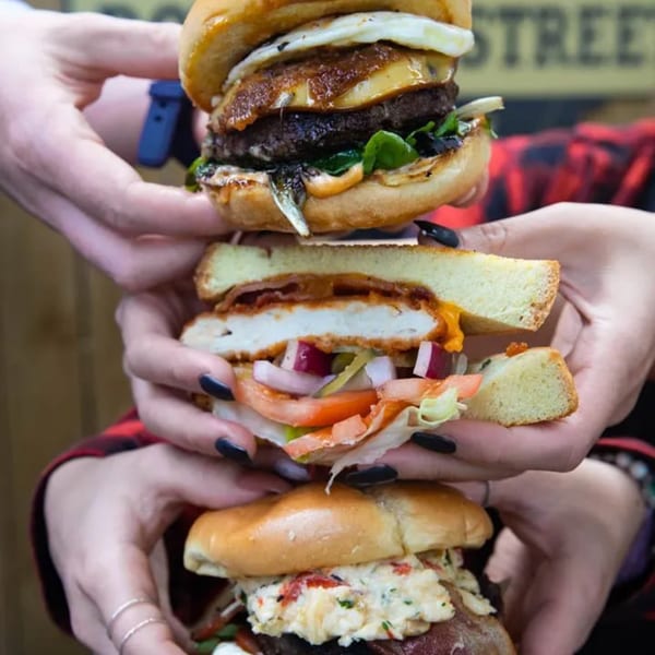 Three hands holding a towering stack of three different gourmet burgers.