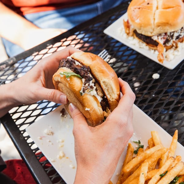 Hands holding a burger with feta cheese and arugula, with fries on a white plate.