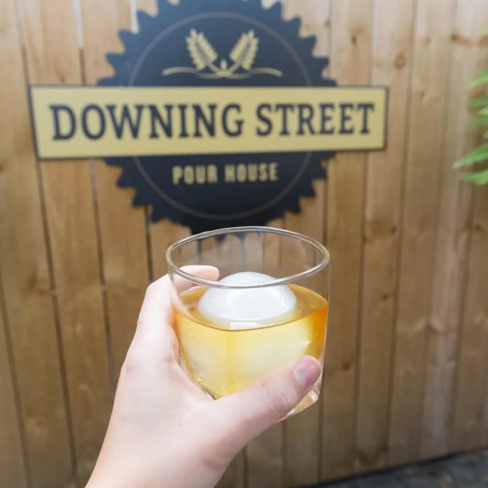 Hand holding a drink with a large ice sphere in front of a wooden fence with a "Downing Street Pour House" sign.