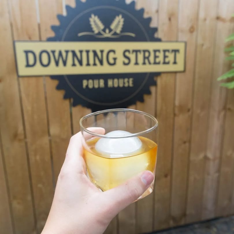 Hand holding a drink with a large ice sphere in front of a wooden fence with a "Downing Street Pour House" sign.