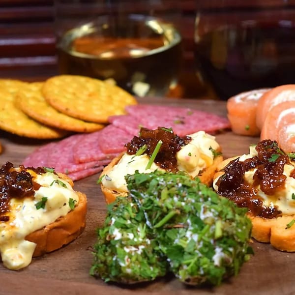 A wooden board with crackers, salami, salmon rolls, and crostini topped with cheese and jam.