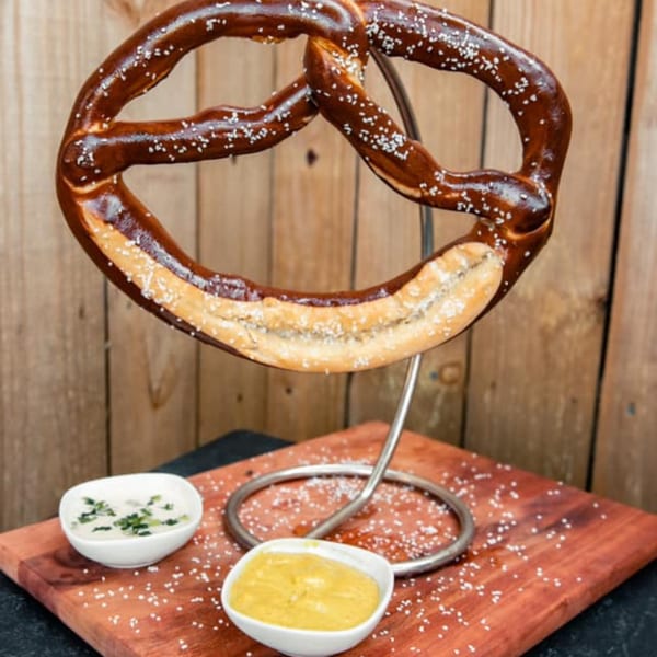 A large, salted pretzel on a metal stand, with two dipping sauces on a wooden board.