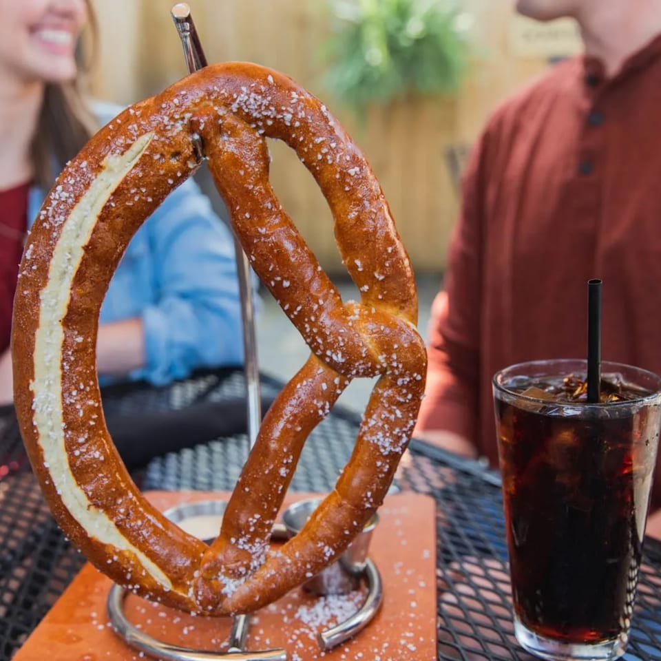 A large, salted pretzel on a stand with dipping sauces, a drink, and people in the background.