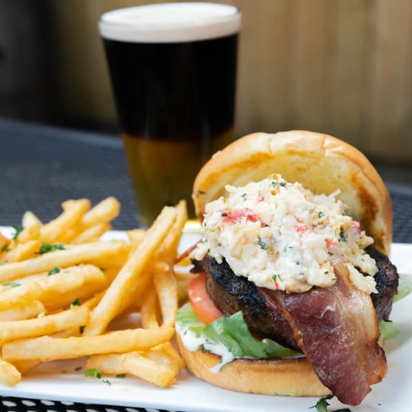 A burger with bacon, lettuce, tomato, and a creamy topping, served with fries and a dark beer.