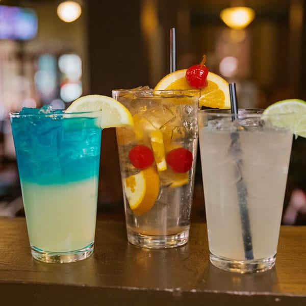 Three colorful cocktails with fruit garnishes and straws sit on a dark wooden bar.